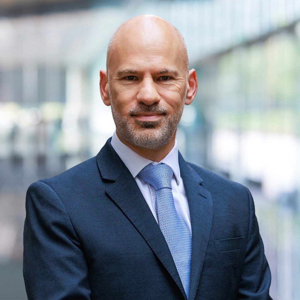 A man in a dark blue suit and blue tie, photographed against a blurred office building background A man in a dark blue suit and blue tie, photographed against a blurred office building background